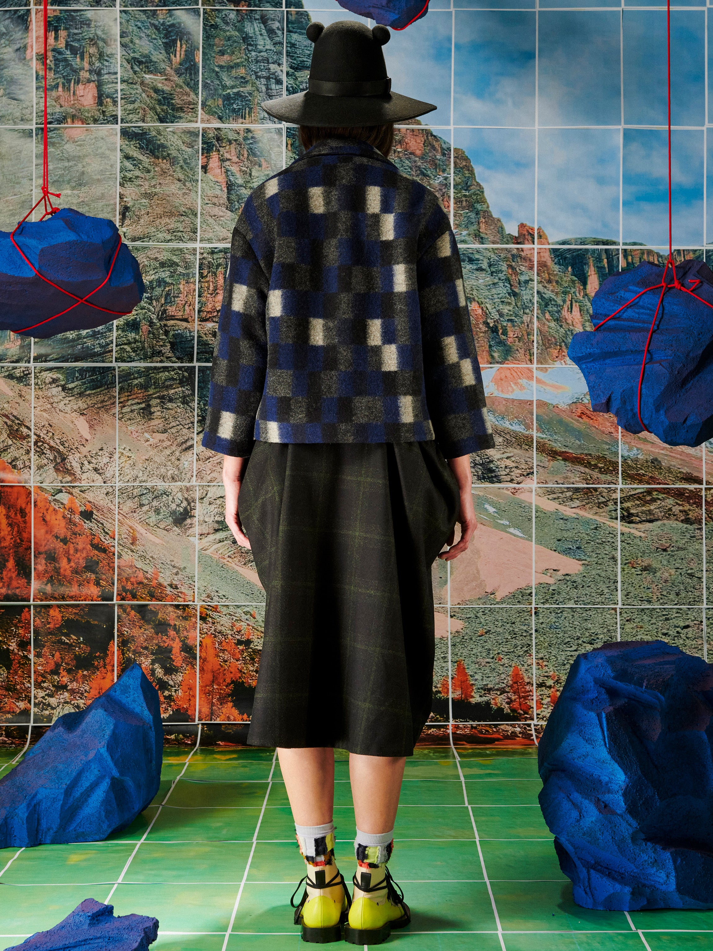 Person wearing a plaid jacket and black hat in front of a tiled wall with blue and red abstract art.