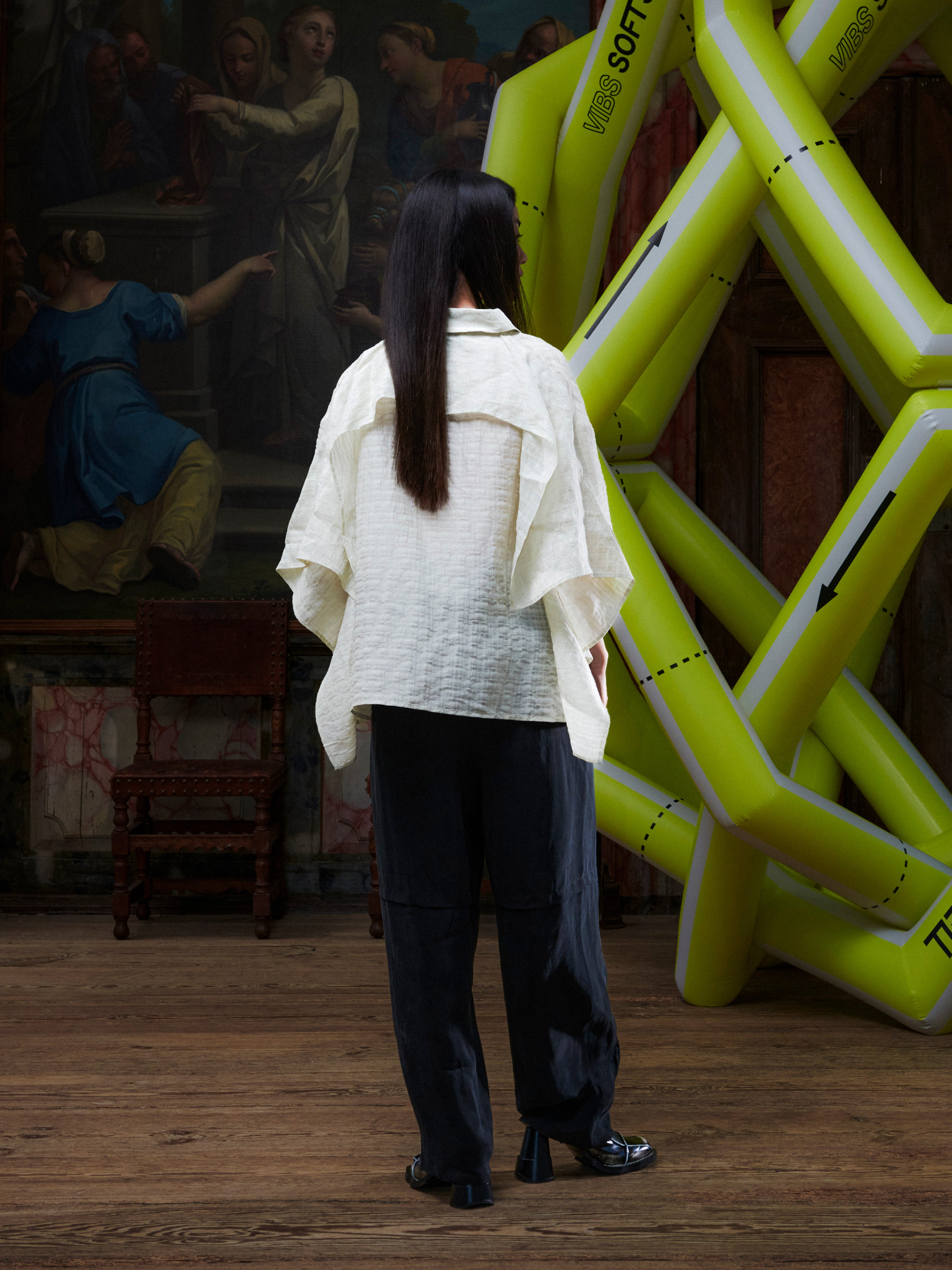 Person wearing a white textured shirt and dark pants standing in front of an inflatable sculpture and artwork.