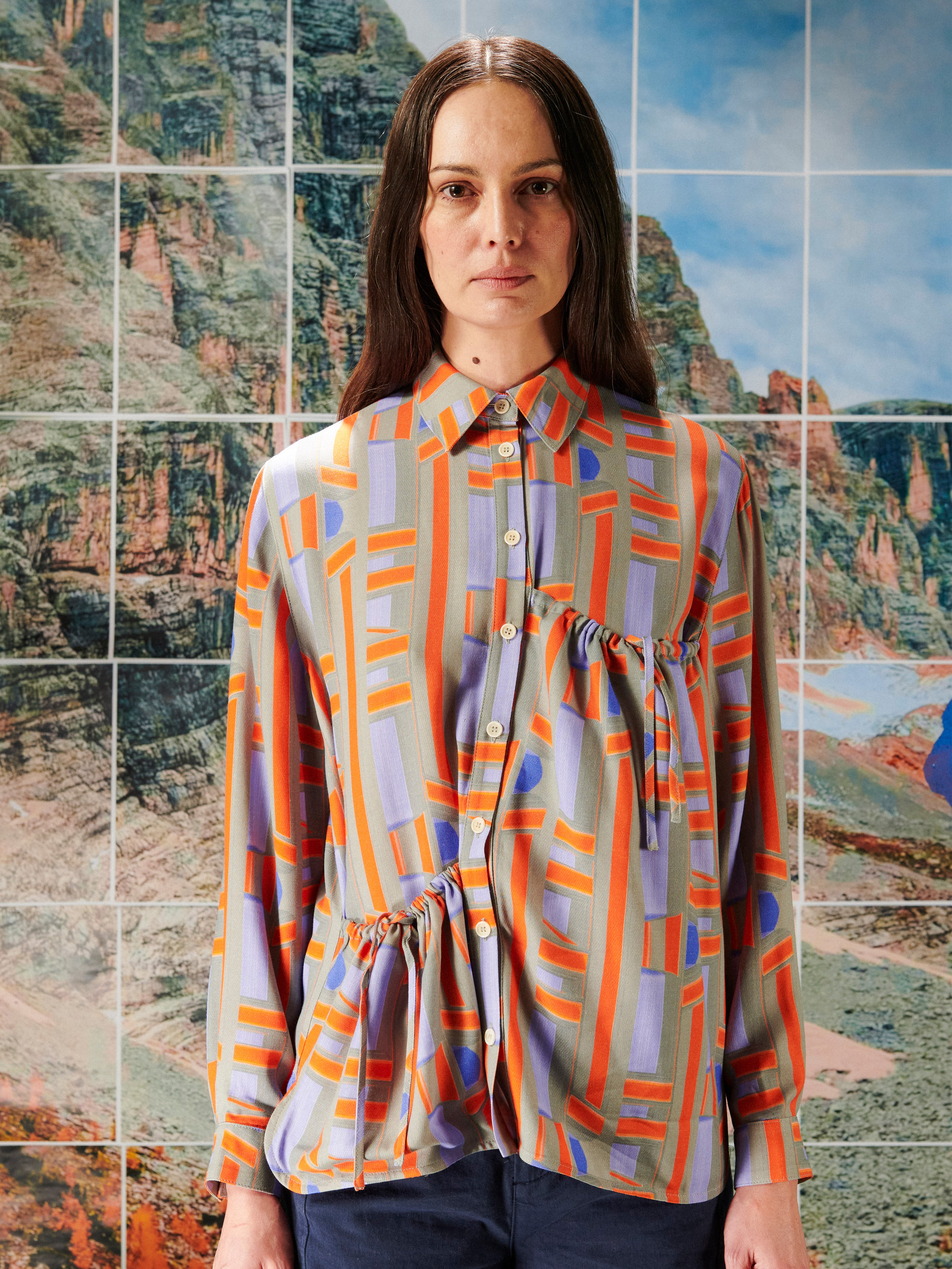 Person wearing a colorful patterned shirt from Henrik Vibskov standing in front of a mosaic of landscape images.