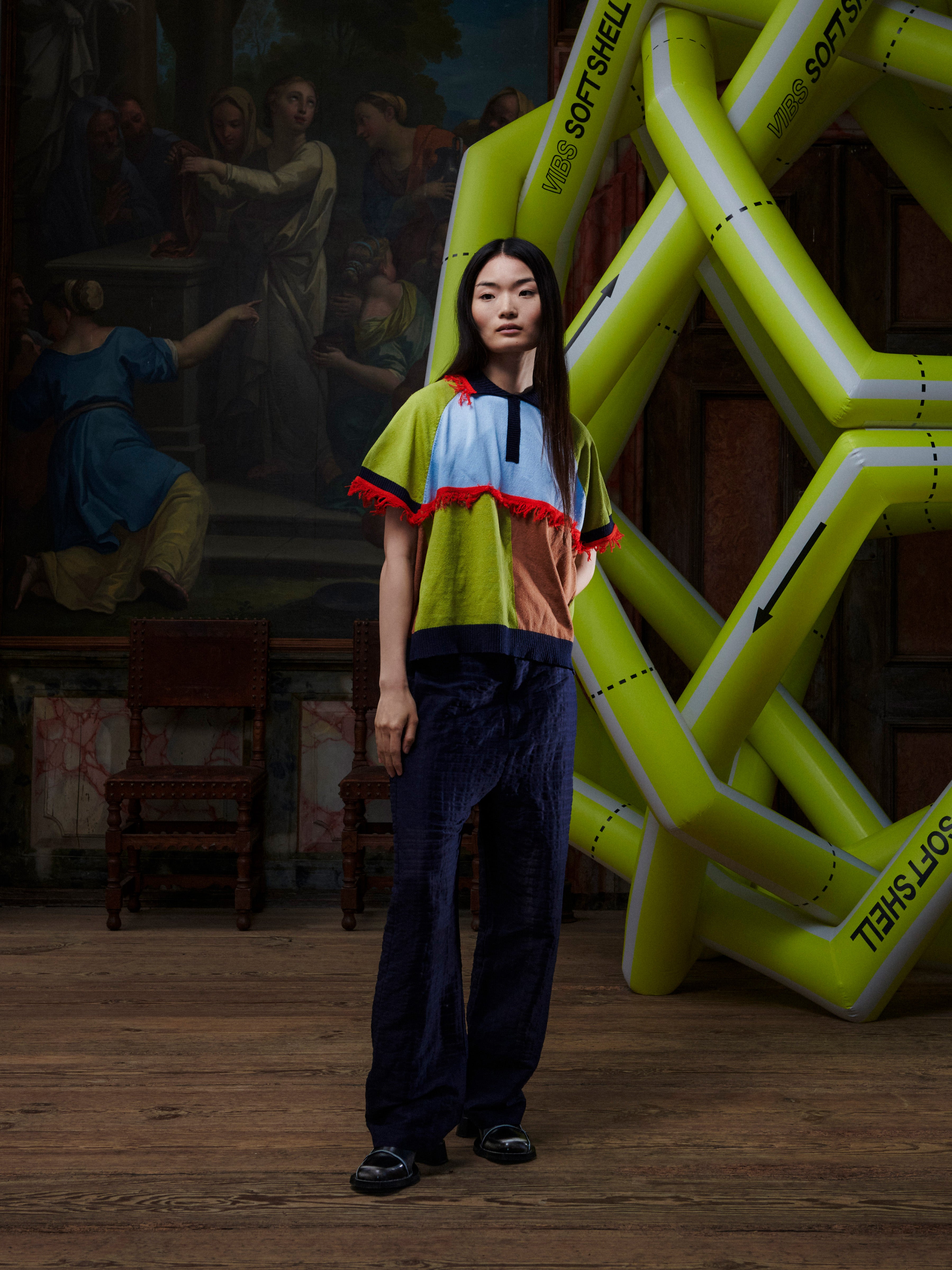 Person wearing a colorful knitted polo with fringe details in an artistic setting with large inflatable letters.