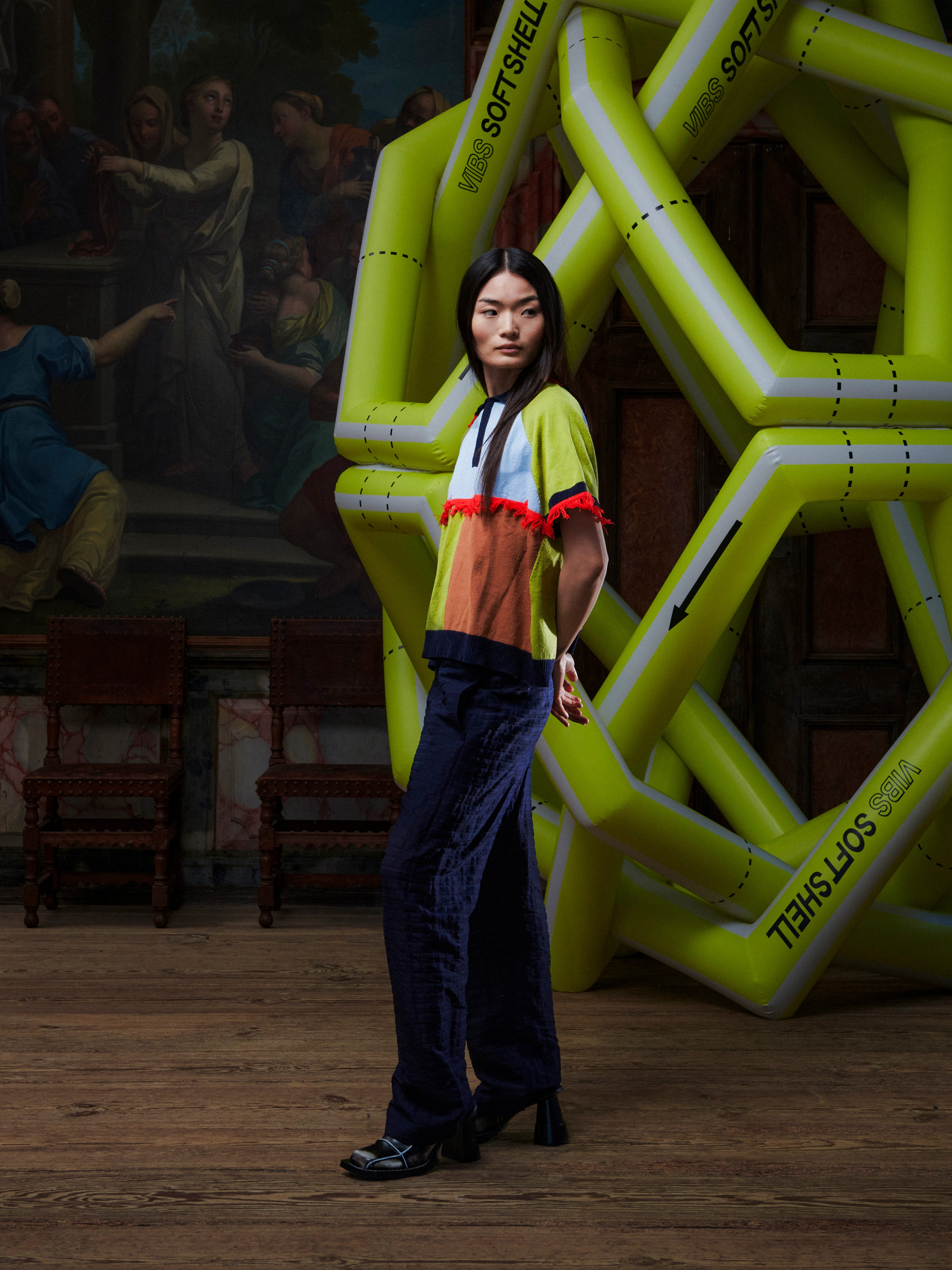 Side profile of person wearing a colorful knitted polo with fringe details in an artistic setting with large inflatable letters.
