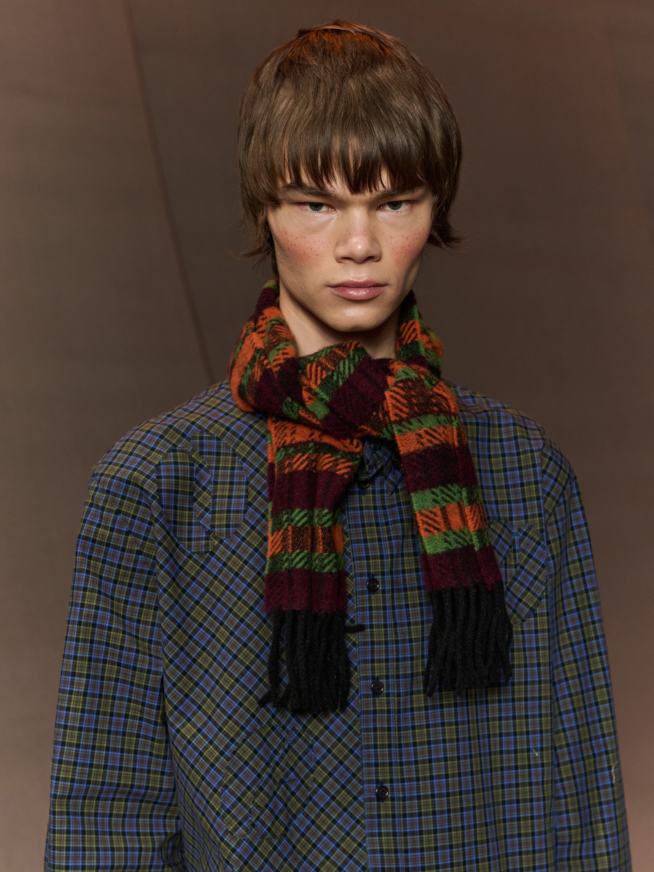Person wearing a plaid shirt and colorful scarf against a brown background