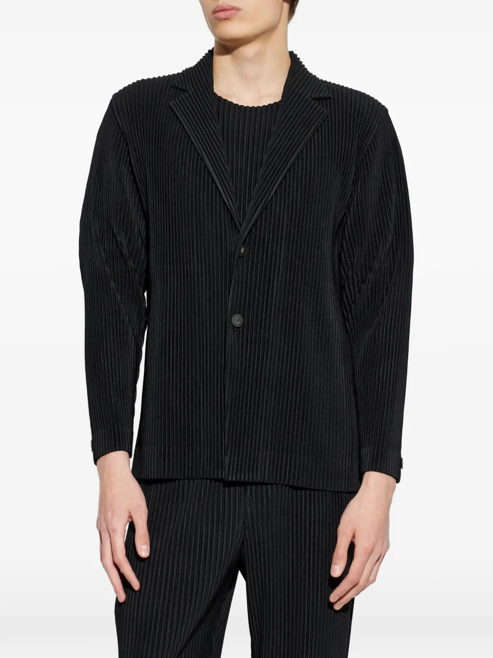 Homme Plissé Issey Miyake April SS26 Basic Jacket black, close-up front view on model showing the structured pleated lapel, V-neckline, and two matte black buttons over a matching pleated top