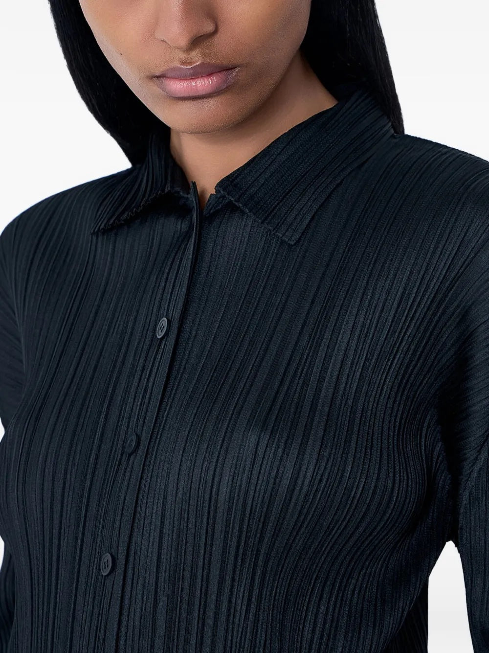 Close-up detail of the model wearing the black, pleated, button-up top with long sleeves from Pleats Please Issey Miyake on a white background