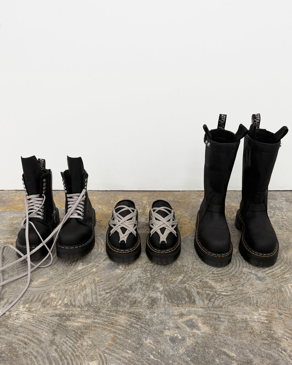 Rick Owens x Dr. Martens FW26 black footwear lineup featuring lace-up ankle boots, low shoes and tall boots with grey laces, pull tabs and yellow welt stitching.
